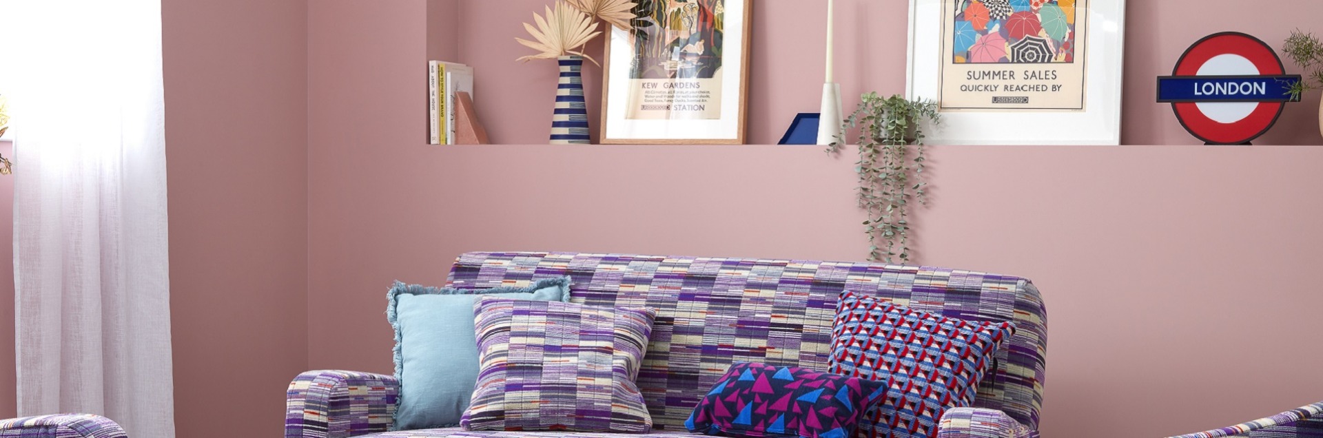 Two framed prints sitting against purple wall with lightbox, with Elizabeth line sofa below featuring scatting of cushions
